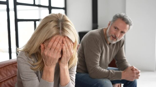 Upset middle-aged couple is arguing at home, frustrated blonde woman covered face with palms and crying, sadness grey-haired mature man on background. Spouses have difficulties, relationship crisis