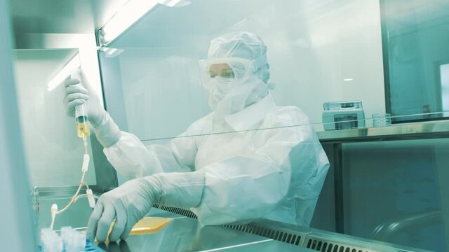 Handheld shot of a female Caucasian scientist in a full protective coverall preparing medical plasma samples in a laminar chamber. Using a syringe to fill the blood plasma container.