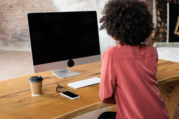 back view of curly african american woman using computer near smartphone, paper cup and eyeglasses...