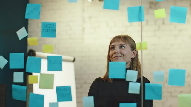 Beautiful Young Caucasian Businesswoman Rearranging Sticky Notes on a Glass Wall Looking Motivated and Focused. Office Employee Wearing a Trutle Neck While Standing in an Industrial Office Space.