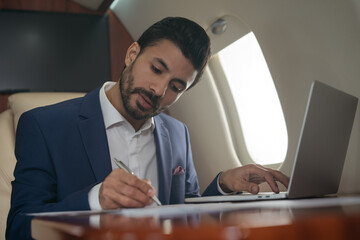 Handsome Arabian businessman using laptop computer, typing, working online sitting in airplane. Confident middle eastern entrepreneur taking notes, signs contract  flying luxury private jet