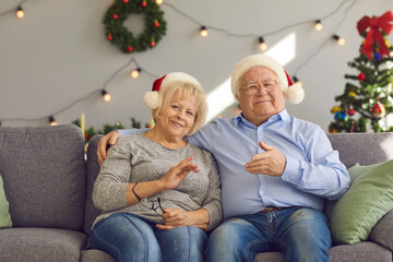 Smiling senior couple in Christmas festive hats sitting on sofa, gesticulating and looking at camera during online videocall with family over decorated room interior. Pensioners celebrating New Year