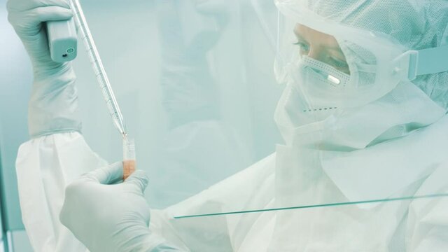 Close-up shot of a Caucasian female lab scientist in a full protective suit, working in a laminar flow cabinet. Using a pipette to put a red liquid in a test tube.