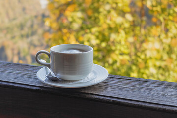 A cup of coffee on a wooden railing against the background of an autumn landscape with sunlight. Blurry autumn background with a hot white cup. White coffee cup on autumn background.