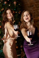 Happy beautiful caucasian women with glasses of wine near Christmas tree. Stylishevening dresses, hairstyle and make up. Festive home interior. New Year's party concept