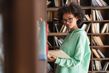 young african american woman in eyeglasses looking at camera while holding book near bookshelf