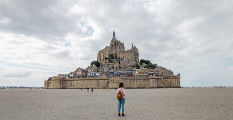 Young woman wearing backpack walking towards mont saint michel with sun behind