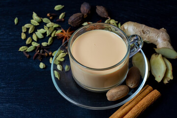 Obraz na płótnie Canvas Masala chai tea in transparent glass cup with ginger, cardamom, cinnamon, nutmeg, and anise on black wooden background. Masala chai popular traditional India hot drink made of milk, tea and spices.