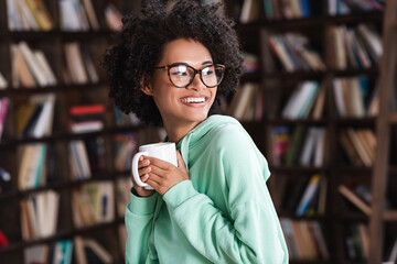 happy young african american woman in eyeglasses holding cup of coffee