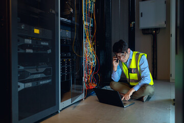 Asian male engineer using laptop and talking on smartphone while inspecting in computer server room - Powered by Adobe