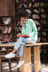 happy african american student in eyeglasses talking on cellphone and reading book while sitting on desk