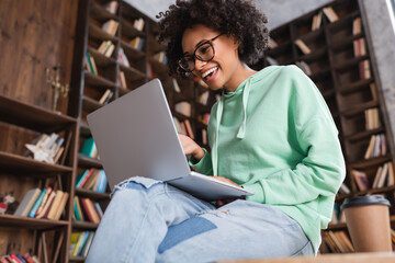 low angle view of smiling african american student in eyeglasses using laptop while sitting on desk