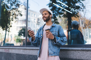 Happy Spanish hipster guy in bluetooth headphones enjoying positive music playlist from mobile radio using 4g wireless in city, cheerful male tourist in earbuds holding coffee to go and smartphone - obrazy, fototapety, plakaty