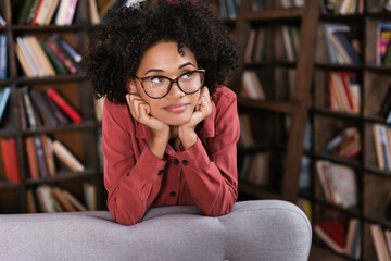 dreamy african american woman in eyeglasses leaning on couch and looking away