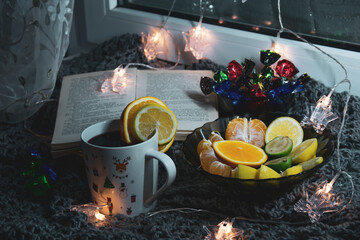 Cozy still life on the windowsill, with an open book, sweets, citrus notes and a garland in the form of asterisks - clothespins. The still life is located on a knitted scarf