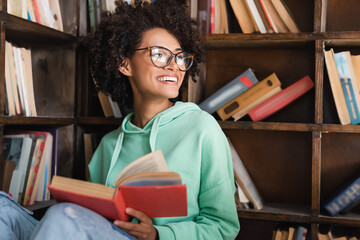 happy african american student in eyeglasses sitting with book in library