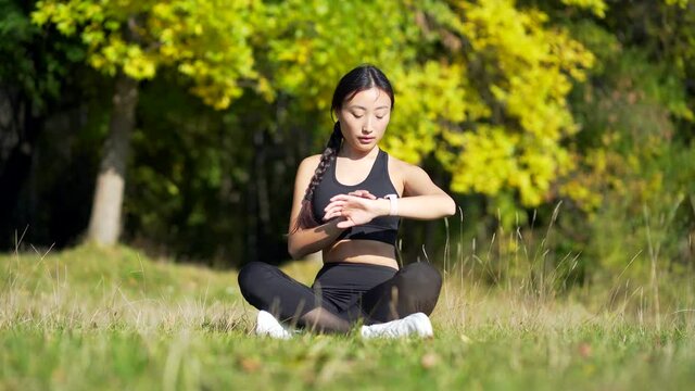 young beautiful sports girl asian woman meditating in park, sitting lotus pose practicing yoga mat outdoors looking smart watch at modern track using use smartwatch fitness bracelet checking result