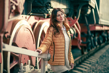 Fototapeta na wymiar A beautiful young woman sits at the wheels of a large vintage train at the station. A woman is resting after a long road, female tourism, rails and sleepers, retro trains. 