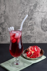 A tall glass of red juice with ice and straws on a gray background, next to a ripe pomegranate fruit, underlaid with a cloth napkin.