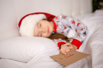 Obraz na płótnie Canvas Merry Christmas and Happy Holidays. The little happy girl sleep on the bed wrote a letter to Santa Claus and holds the envelope in her hand.