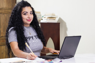 Fototapeta na wymiar Latin girl with curly hair studying her university degree from the comfort of her home on her laptop while listening to music with her gaming headphones
