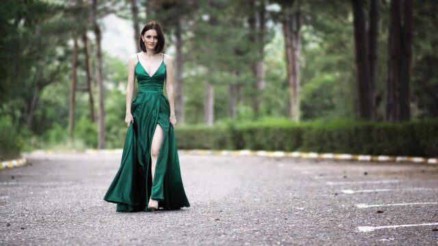 Beautiful smiling young woman in a long luxury silk satin green dress with long waving train walking on the empty road during a photo shoot.