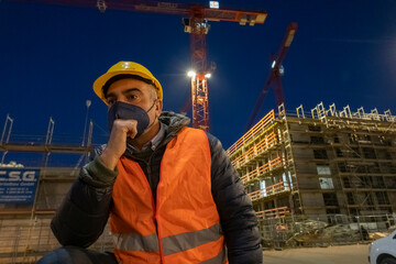 Engineering during COVID-19 Coronavirus outbreak. Occupational safety and health and protection. Construction worker posing at night wearing safety vest, hardhat and medical mask