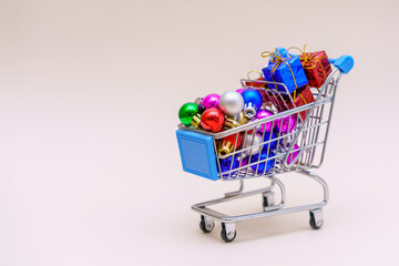 Shopping cart with multicolored christmas balls for decorating christmas tree on a light pink background. Basket with creative christmas gifts. Christmas decoration concept