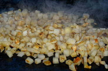 Hash browns being cooked on a griddle, with steam rising from the small cubed potatoes