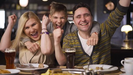 Front view excited Caucasian family gesturing victory smiling sitting in restaurant indoors. Portrait of happy man woman and teenage boy rejoicing winning watching sport match. Slow motion