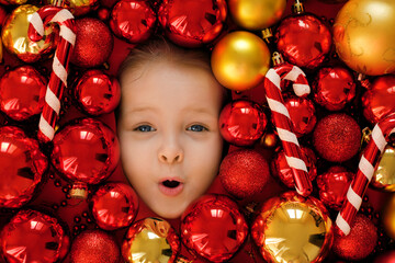 The surprised, enthusiastic, pretty face of a child in a heap of bright red and gold Christmas balls. Template for New Year, winter holidays promotions, discounts and sales with copy space