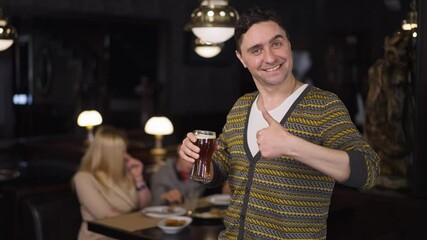 Portrait of happy man with pint of beer gesturing thumb up looking at camera smiling. Positive confident Caucasian father husband dining with family in restaurant advertising alcohol. Slow motion