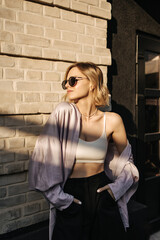 Photo of sexy caucasian young blonde standing against brick wall in sunlight. Girl in sunglasses holds her hands in pockets of her trousers and looks to side.