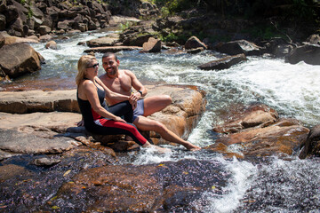 happy couple talking and relaxing, sitting on rock with river water in sunny day.