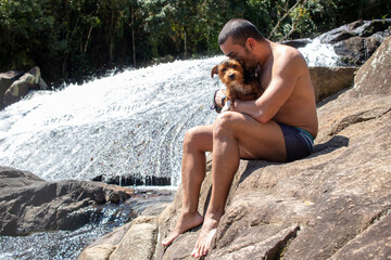 man sitting on stone kissing his little black dog, admiring the waterfall in sunny day.