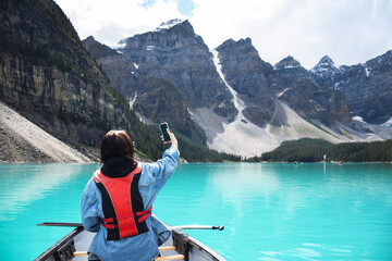 Young girl taking selfie with mobile phone while riding in canoe on turquoise lake with view of mountains