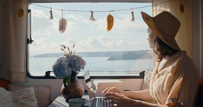 Young woman wearing hat in campervan typing on laptop with beatiful ocean view and lensflares shot in 4k super slow motion