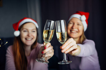 Christmas toast with glasses of champagne in hands. Blurred background: two girls in Santa Claus hats, clink glasses and wish Happy New Year. Love, friendship, family.