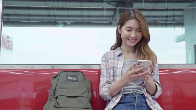 Young Asian woman sitting on train to travel, smiling and chatting with friend on smartphone.