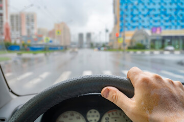 the driver hand on the steering wheel inside the car against the background of the streets of an empty city without people, city buildings, zebra crossing in cloudy, dark, rainy weather