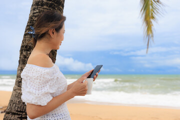 Woman holding coffee cup and smartphone standing under coconut palm trees