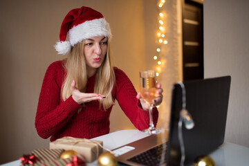 Portrait of happy girl with blond hair holding hand blowing kisses and glass of champagne looking at laptop, online greeting merry christmas or happy new year