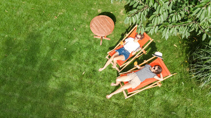 Young girls relax in summer garden in sunbed deckchairs on grass, women friends have fun in green...