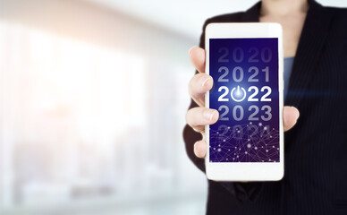 Success new year concept. Business management, Inspiration concepts ideas. Hand hold white smartphone with digital hologram 2022 sign on light blurred background.