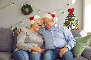 Smiling loving elderly couple in Christmas festive caps sitting on sofa and hugging during Christmas over decorated room interior. Pensioner couple celebrating New Year holidays together at home
