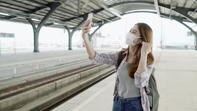 Young Asian woman backpacker wearing medical mask, using smartphone to taking selfie on platform at red line train station.