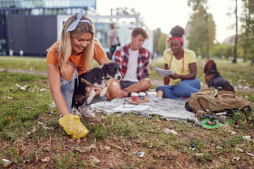 A young girl is picking up the poop of her dog while having a good time with her friends in the park. Friendship, rest, pets, picnic