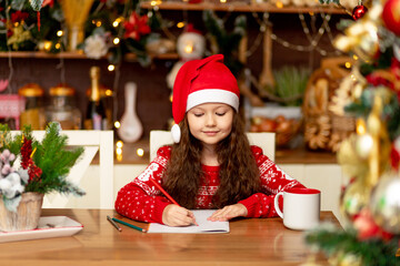a cute little girl child in winter clothes a Santa Claus hat and a red sweater writes a letter to Santa Claus in the dark kitchen of the house and smiles and waits for the new year and Christmas
