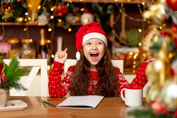 a cute little girl child in winter clothes a Santa Claus hat and a red sweater writes a letter to Santa Claus in the dark kitchen of the house and smiles and waits for the new year and Christmas