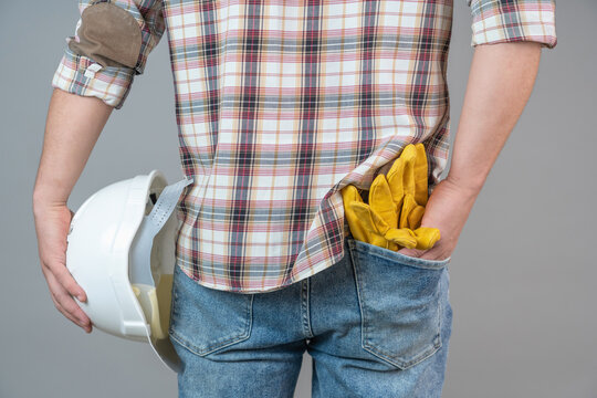 Close-up of a man from behind holding a protective helmet and work gloves in the back pocket of his pants on a gray background.